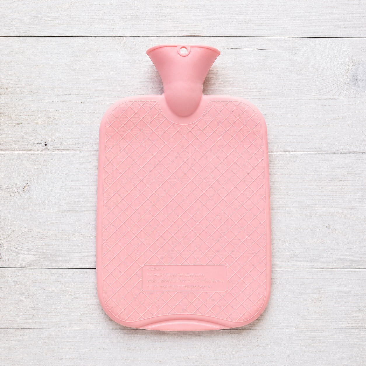 A pink hot water bottle with a diamond-textured cover and a matching pink neck, laid flat on a white wooden plank background.