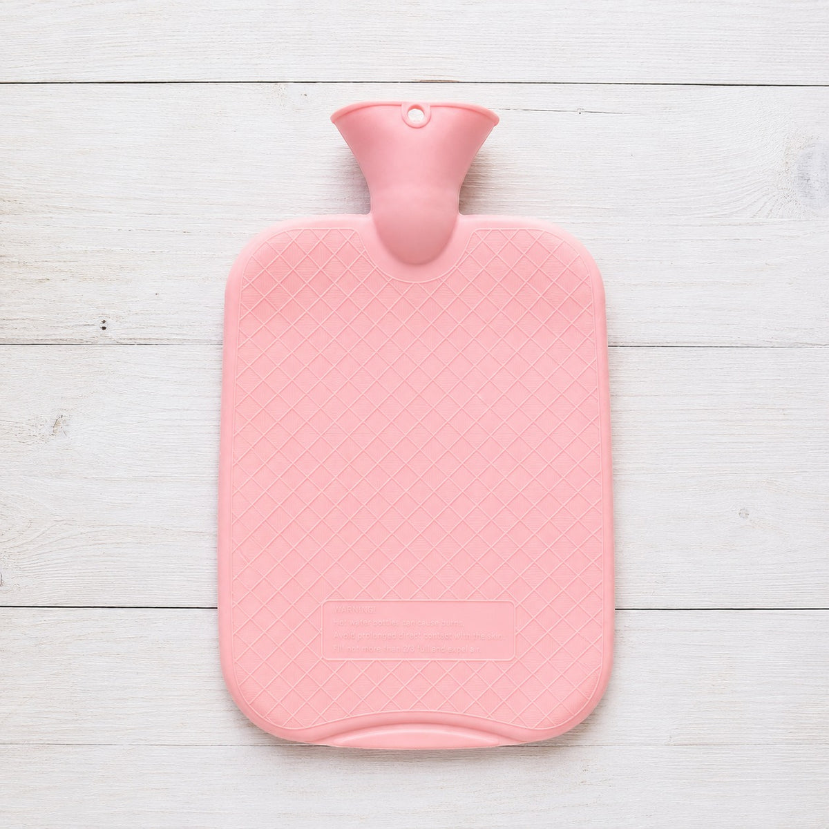 A pink hot water bottle with a diamond-textured cover and a matching pink neck, laid flat on a white wooden plank background.