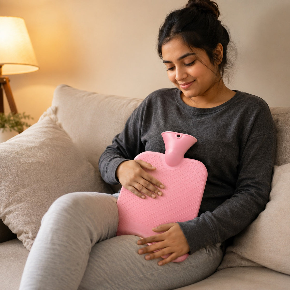 A woman sitting on a beige couch holds a pink hot water bottle against her stomach in a cozy, warmly lit living room setting.