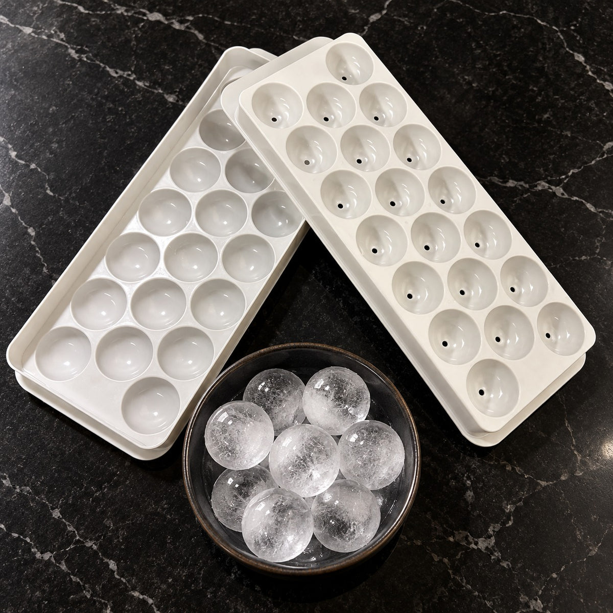 Two white silicone ice cube trays with round domed cavities are shown on a dark marble surface, with a small bowl of clear spherical ice cubes placed in front.