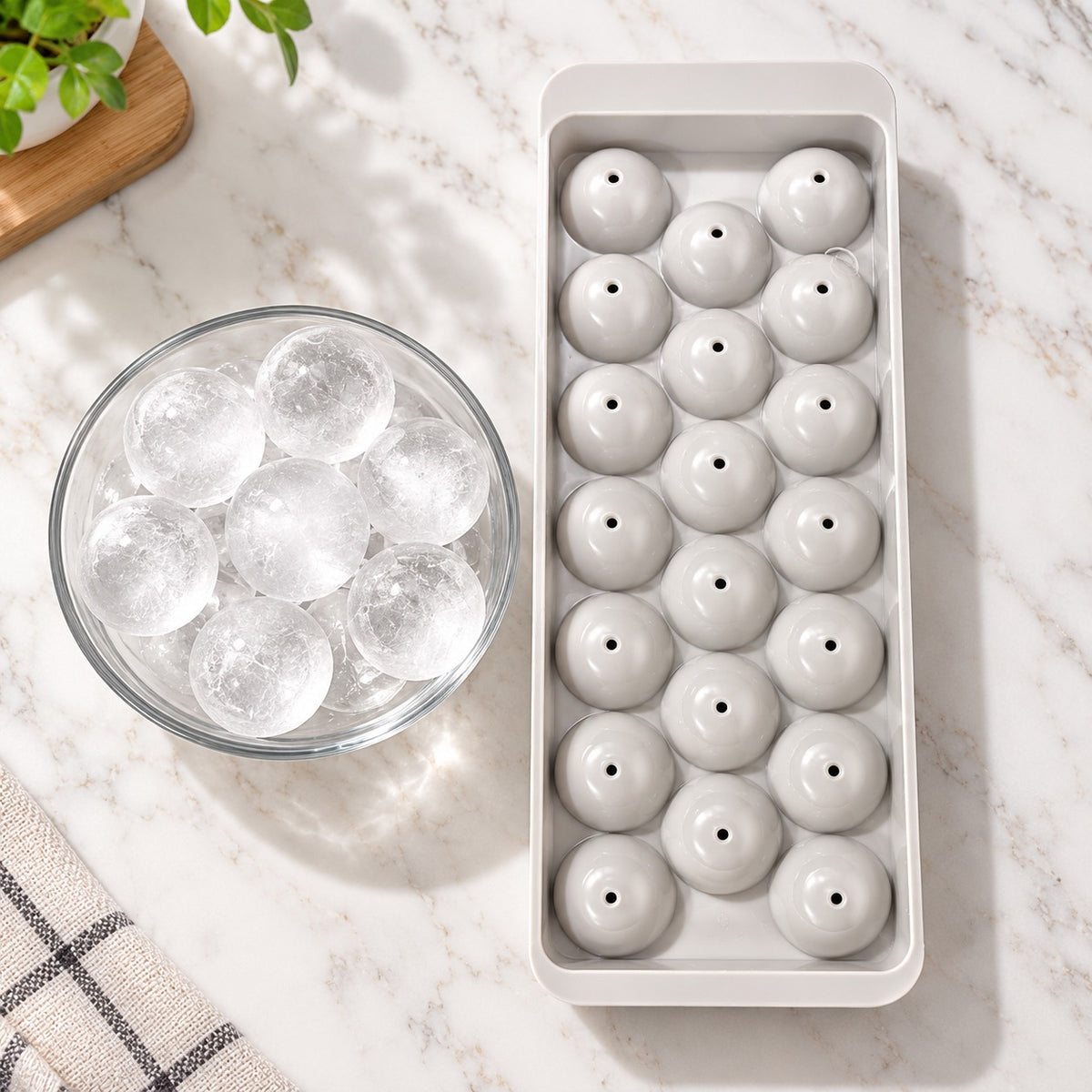 A rectangular ice mold tray filled with round ice spheres, shown beside a glass bowl of clear ice on a white marble surface with a small plant and cloth visible in the background.