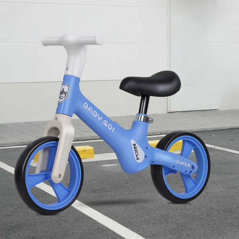A blue and white balance bike with a black saddle and black tires, parked on a gray paved surface in front of a white wall.