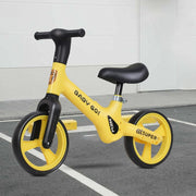 A yellow and black balance bike with 'BABY GO!' and 'SUPER' branding, photographed on a gray paved surface against a light gray wall.