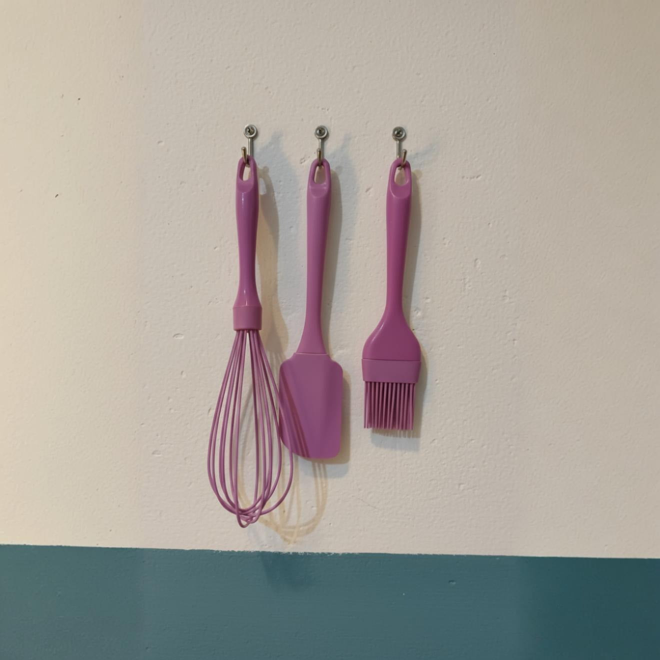 A whisk, a spatula, and a basting brush are hanging neatly on hooks against a wall.