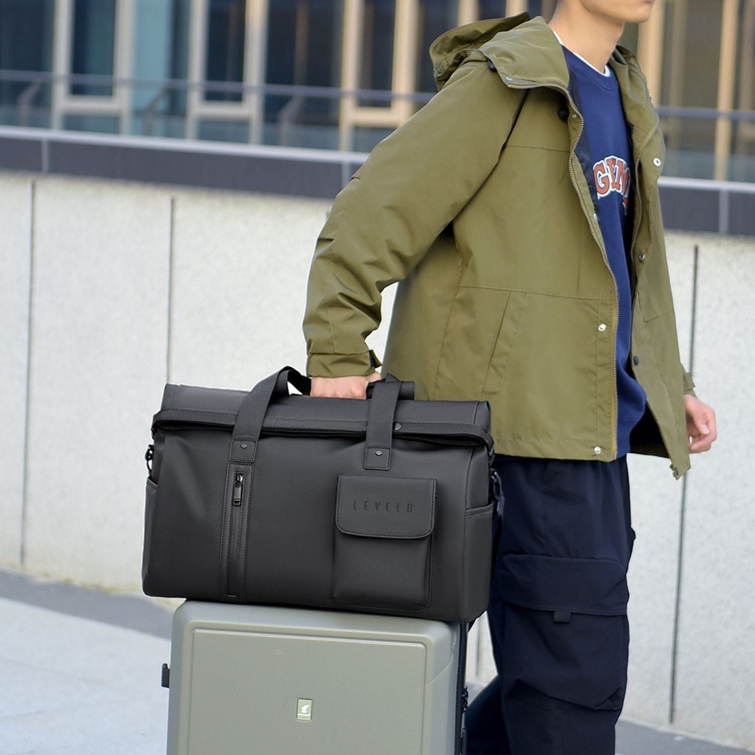 A man wearing an olive green hooded jacket and navy pants carries a LEVELO Monaco Waterproof Travel Duffel Bag on his shoulder while standing next to a gray suitcase outdoors.