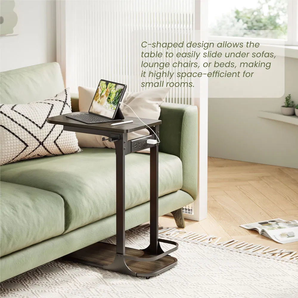 A dark brown C-shaped adjustable laptop table placed next to a light green sofa in a bright living room. A laptop is open on the tabletop, and a text overlay explains the table's space-saving C-shaped design.