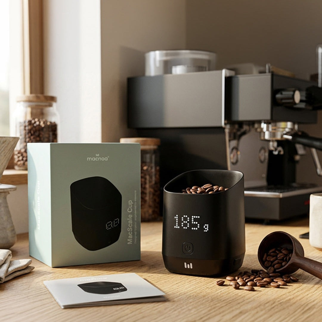 Macnoa MacScale Cup Digital Coffee Dosing Cup with a box on a wooden surface, with coffee beans and a coffee machine in the background.