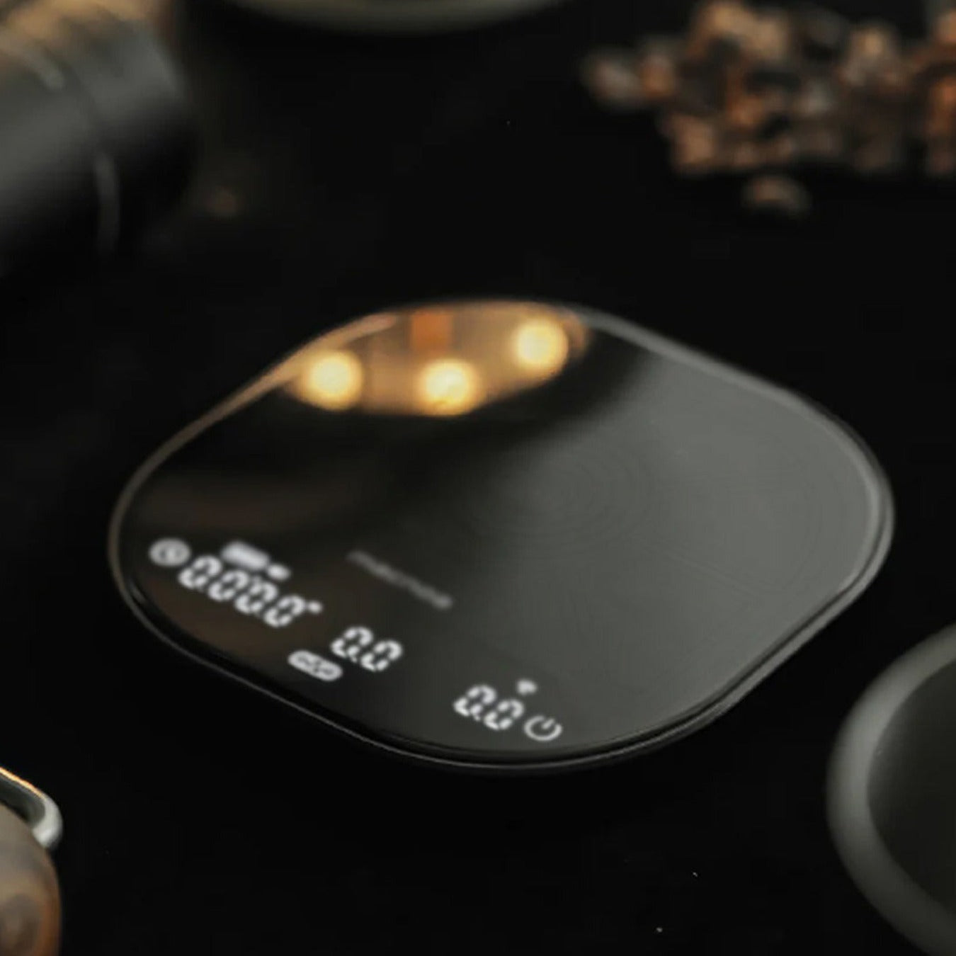 A sleek Macnoa MacScale Pro Smart Coffee Scale with a glossy finish and illuminated white LCD display, shown on a dark tabletop with softly blurred coffee beans and kitchen items in the background.