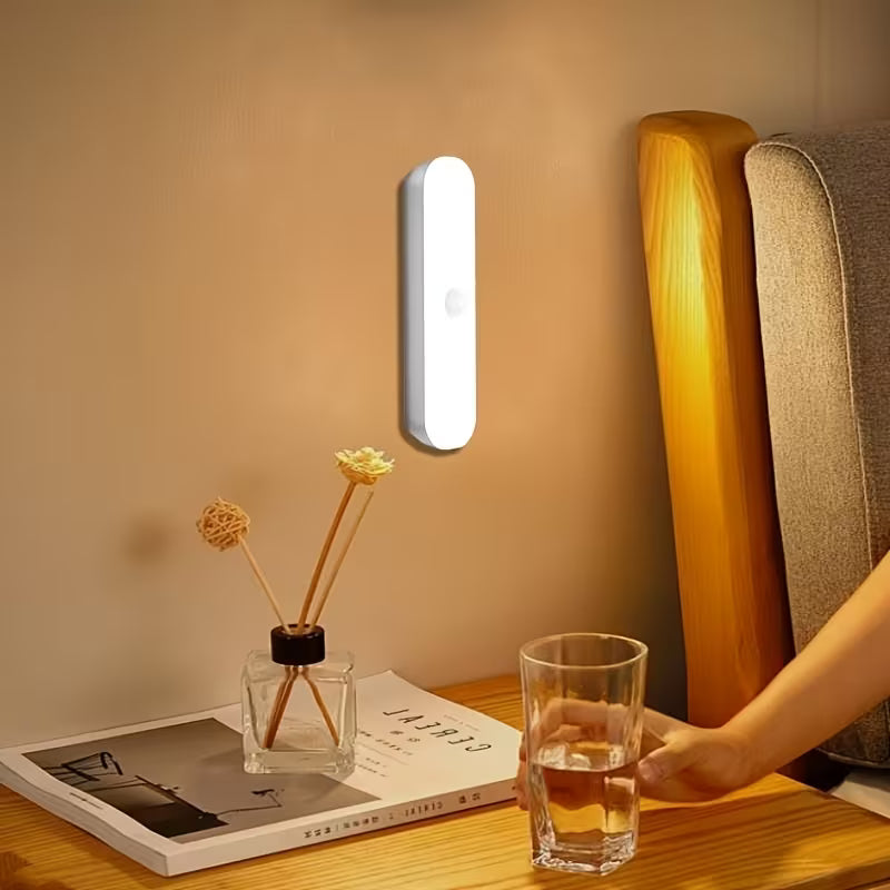 A slim white motion sensor night light mounted on a bedroom wall, softly illuminating a bedside table with a glass of water, reed diffuser, and book, creating a warm nighttime atmosphere.