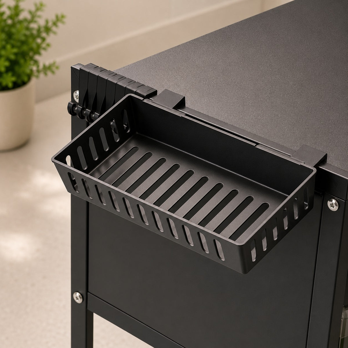 A black metal storage basket with a slatted front, mounted on the side of a Over-Sink Dish Drying Rack in a clean indoor setting.