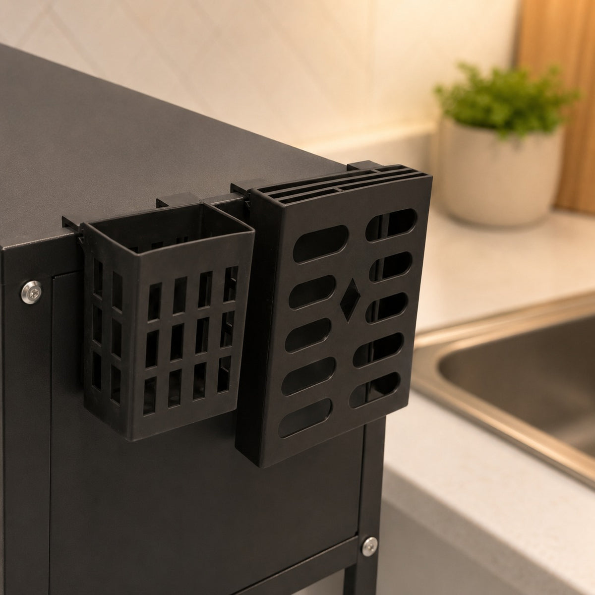 A black metal kitchen organizer rack mounted on the side of a Over-Sink Dish Drying Rack, with a sink and potted plant blurred in the background.