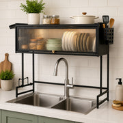 A black metal kitchen sink organizer with a glass-front upper cabinet, shown installed over a double-basin stainless steel sink in a bright kitchen. The organizer holds plates, bowls, and kitchen items, with a faucet and tiled backsplash behind it.