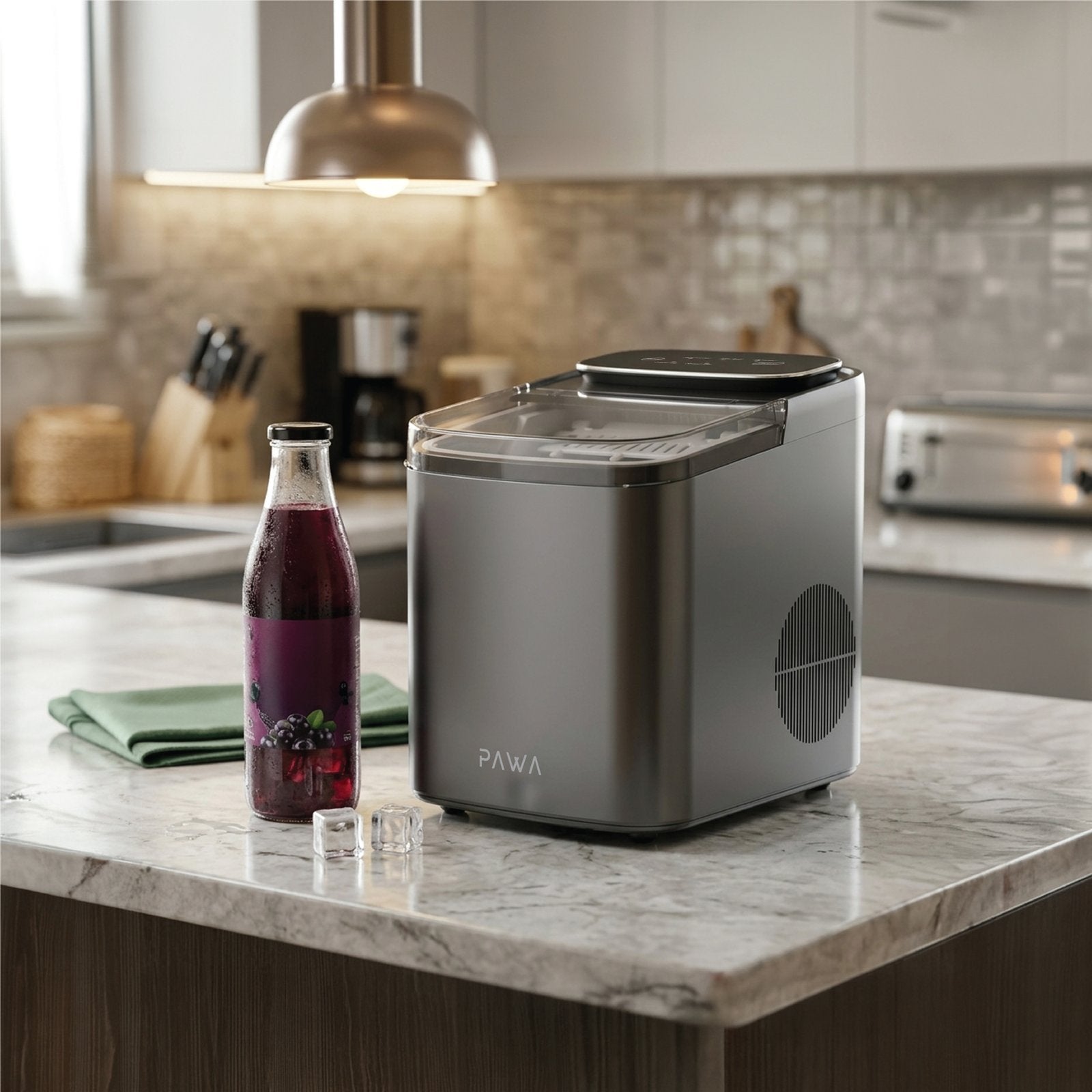 Pawa Chiller Portable Ice Maker in a brushed silver finish sits on a marble kitchen island next to a glass bottle of dark berry drink, with a folded green cloth and small ice cubes in a blurred modern kitchen background.