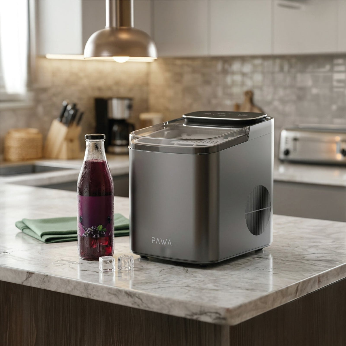 Pawa Chiller Portable Ice Maker in a brushed silver finish sits on a marble kitchen island next to a glass bottle of dark berry drink, with a folded green cloth and small ice cubes in a blurred modern kitchen background.