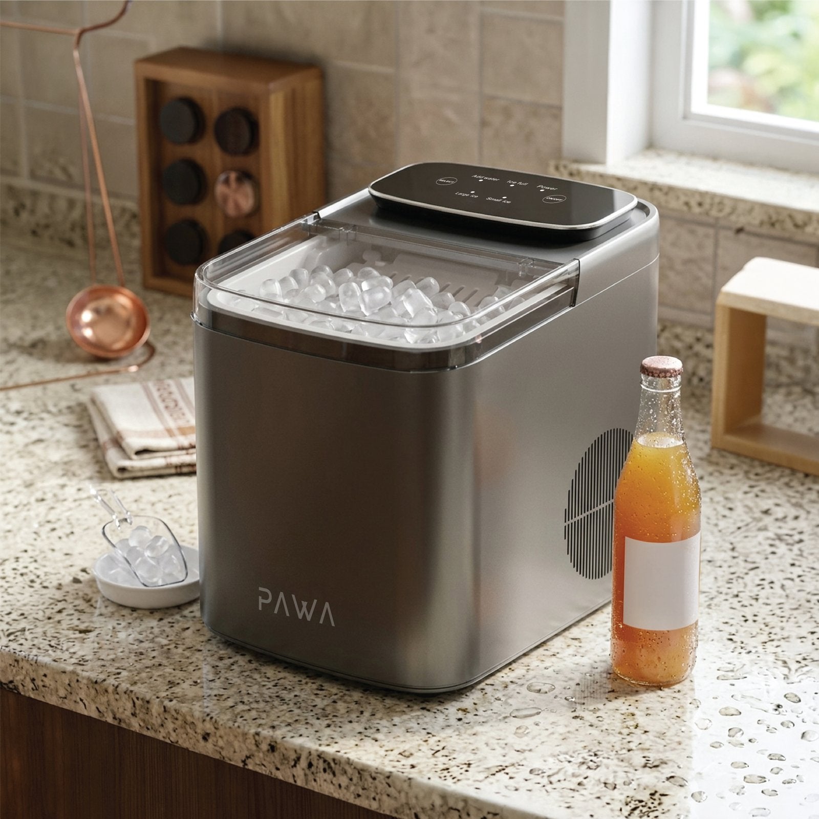 Pawa Chiller Portable Ice Maker in brushed stainless steel sits on a speckled kitchen counter, filled with ice cubes. A glass bottle of orange drink and a small ice scoop are placed beside it, with a tiled wall and window in the background.