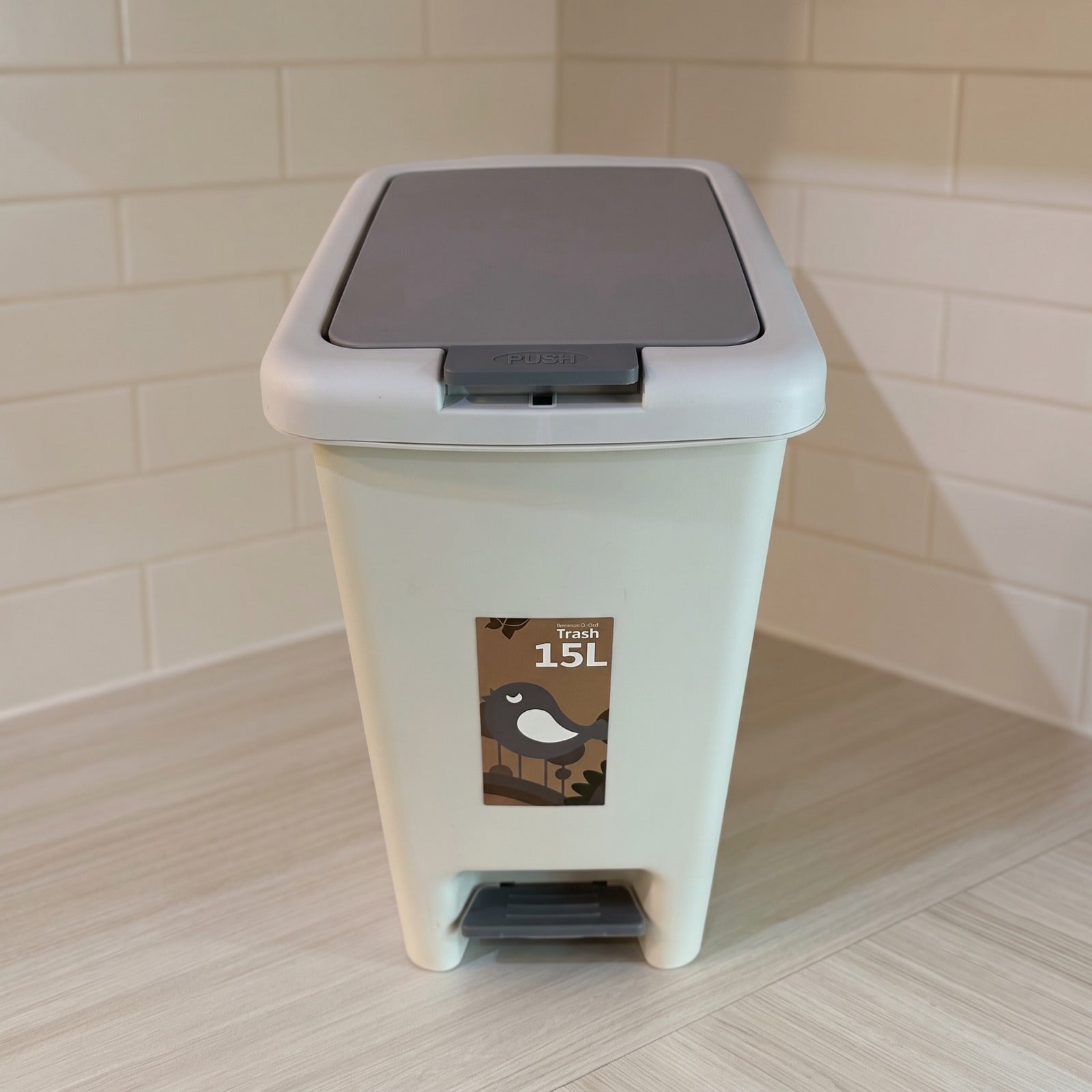 A 15L plastic trash can with a light beige body, a dark gray flip-top lid, and a foot pedal base, set against a tiled wall and light wood floor.