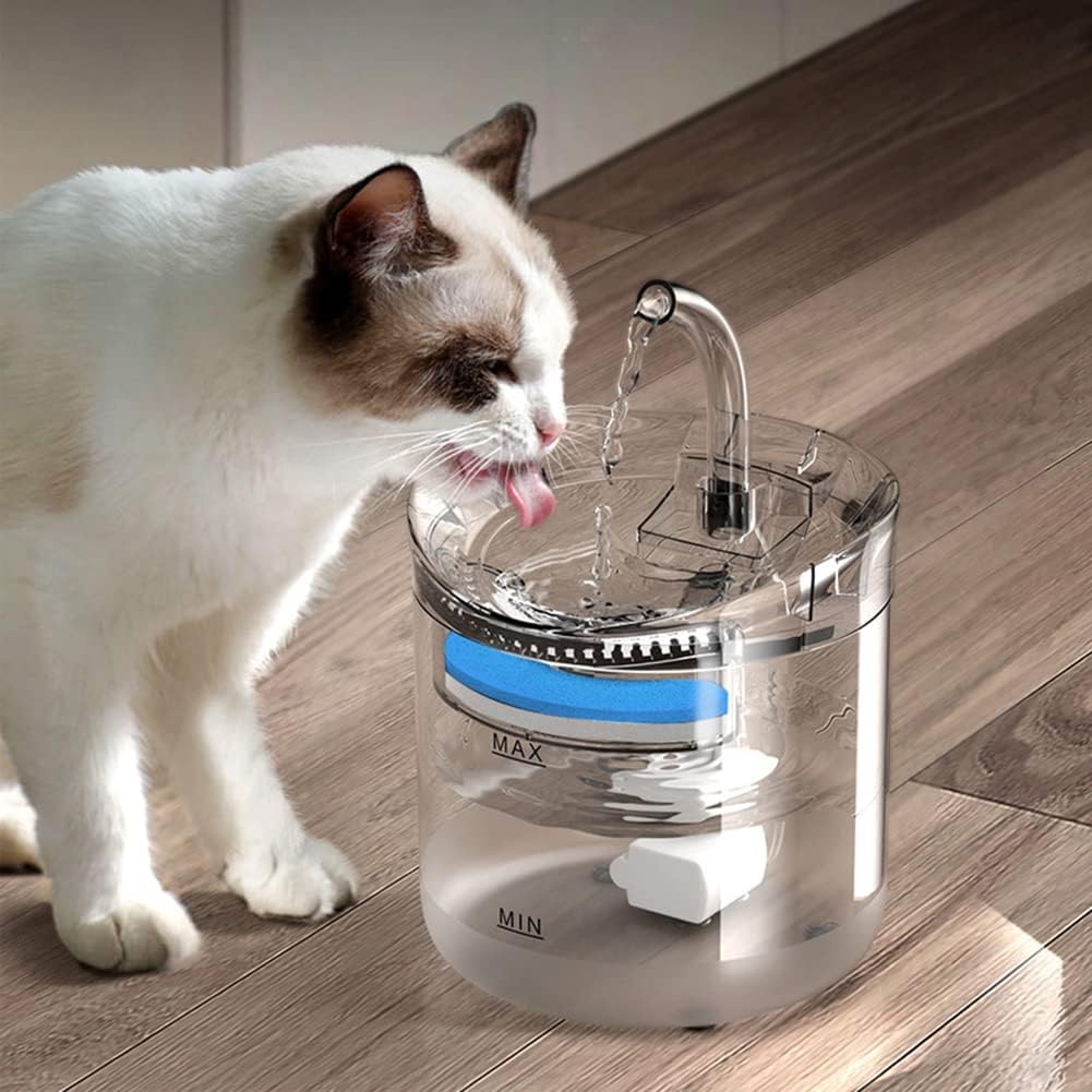A cat drinking water from a transparent pet water fountain placed on a wooden floor, with a continuous stream flowing from a spout, showing the filtered water system and fill level markings.