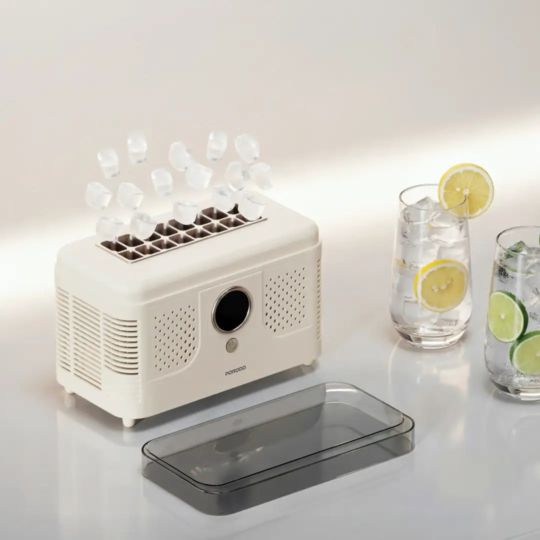 A compact PORODO FrostIQ Portable Ice Cube Maker with a transparent lid placed in front, shown on a clean light background. Several clear ice cubes are visible above the top tray, and two glass drinks with lemon and lime slices sit to the right as styling props.