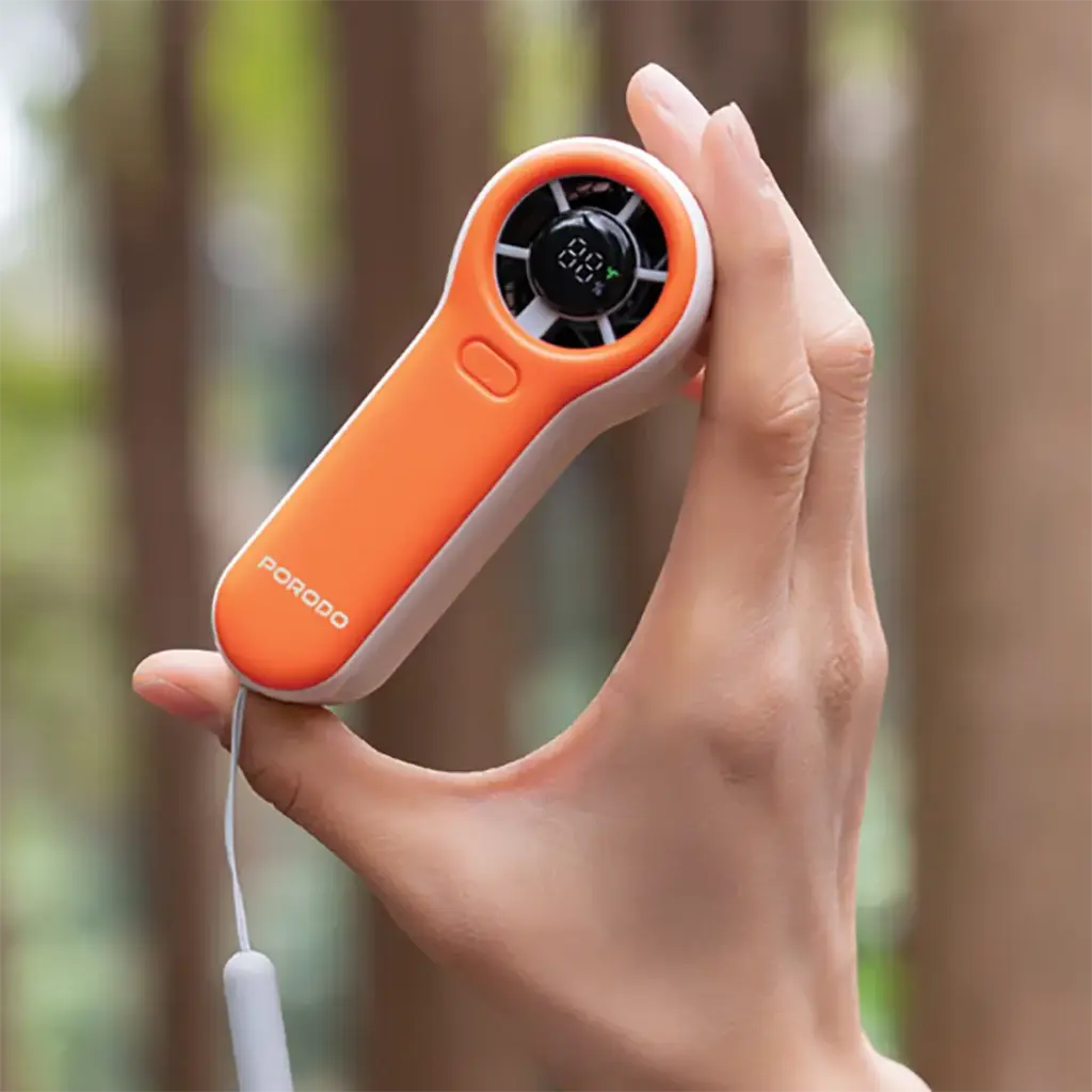 Hand holding an orange Porodo Pocket Size Handheld Fan with a blurred natural background