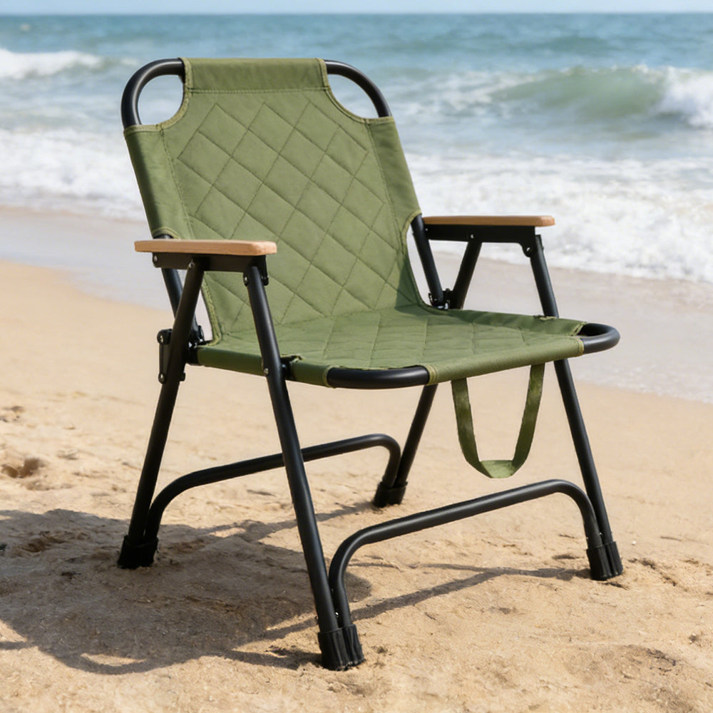 A Folding Camping Chair with a black metal frame and light wood armrests, set on a sandy beach with the ocean waves in the background.
