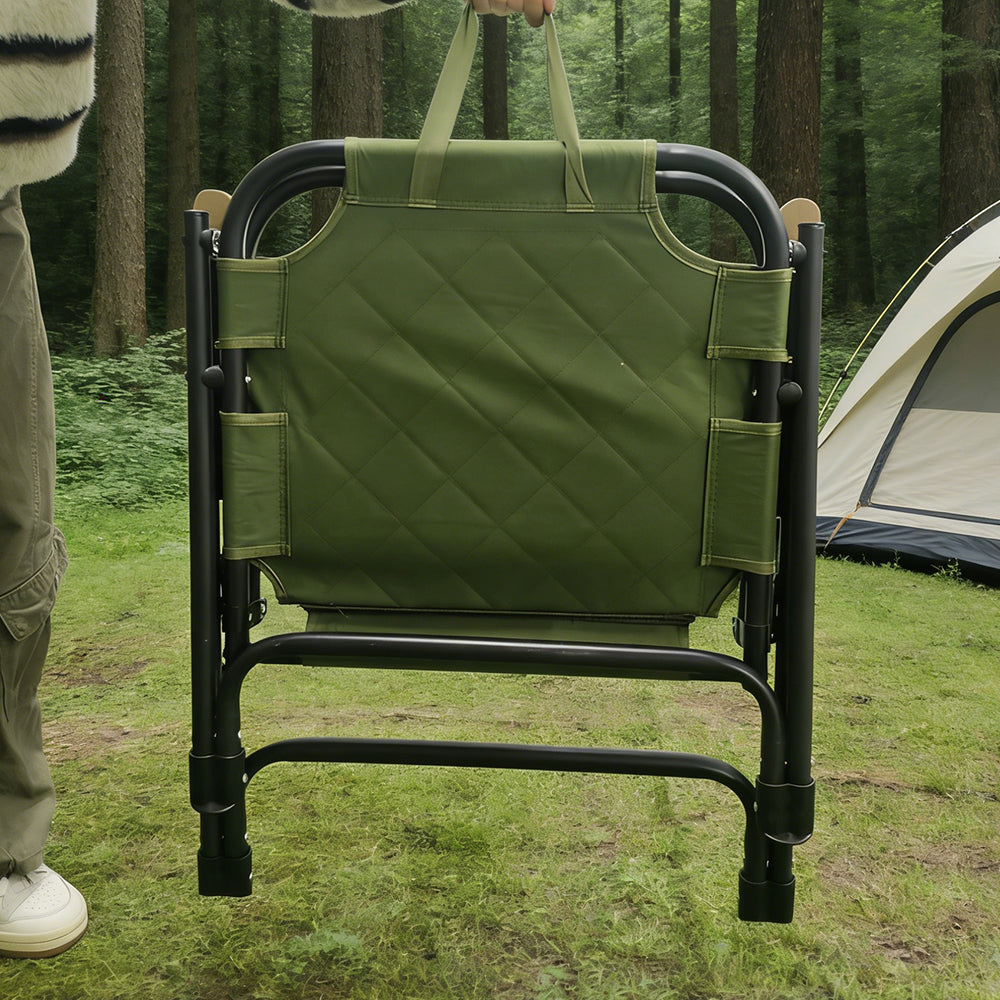 An olive green padded camping chair with a black metal frame, being held by a person in a forest setting with a tent visible in the background.