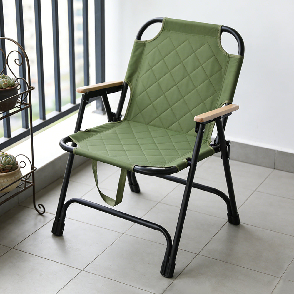 A Portable Folding Camping Chair  with a quilted olive green fabric seat and backrest, light wood armrests, and a black metal frame, positioned on a tiled balcony.