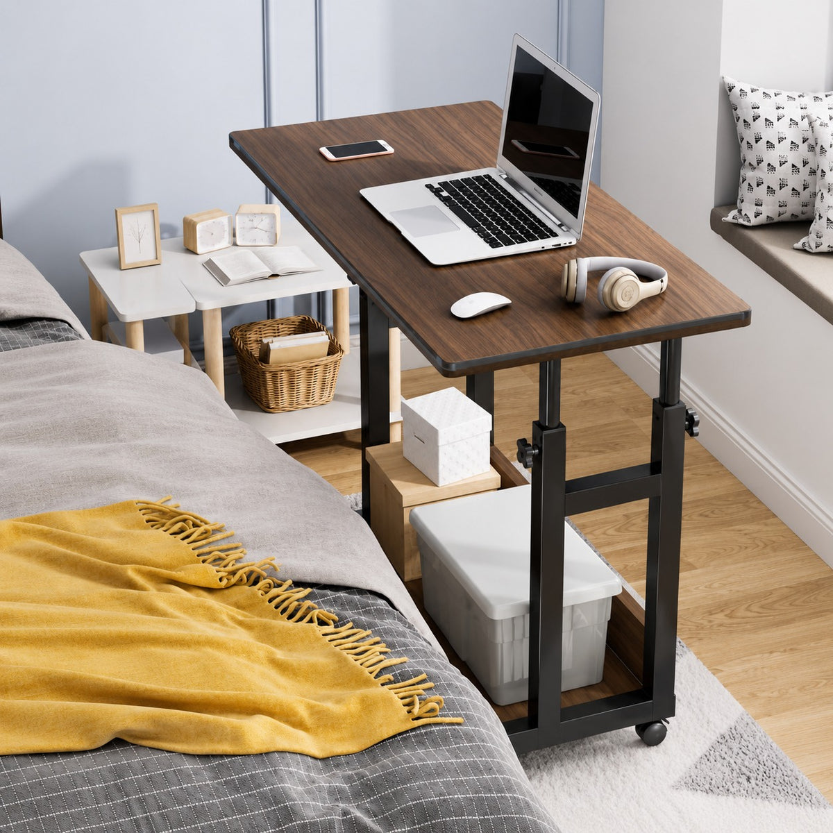 Wooden adjustable desk next to a bed with a laptop and other items on it.