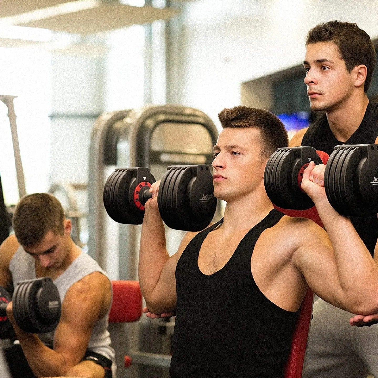 Three men working out in a gym. Two of them are lifting adjustable dumbbells, and one is assisting or spotting