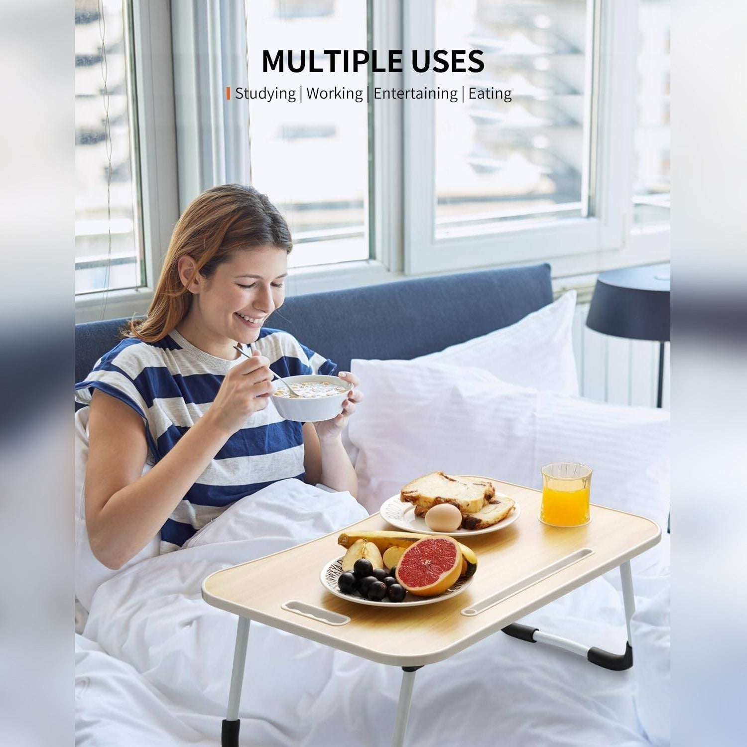 A woman in a blue and white striped shirt sits in bed using a light Foldable Laptop Table  with a tray table setup. The scene includes a bowl of cereal, a plate of toast and fruit, and an orange juice glass, with the text 'MULTIPLE USES' at the top.
