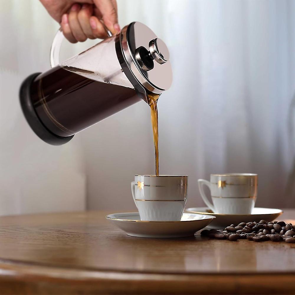 A hand pouring dark coffee from a clear glass French Press Coffee Maker into a glass mug on a white surface.