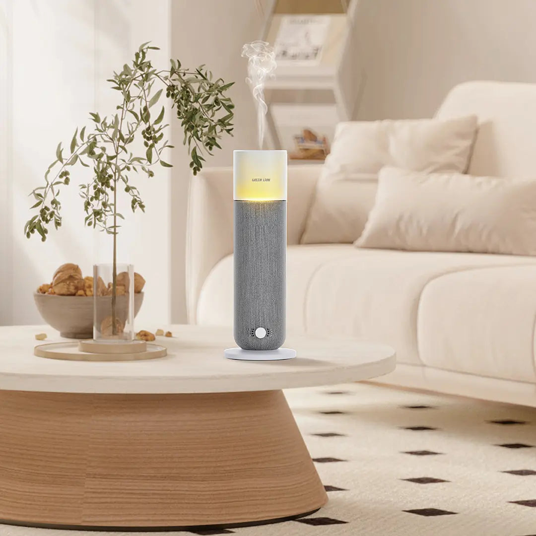 The Green Lion Smart Bloom Aroma Diffuser is placed on a light-colored circular wooden coffee table in a cozy living room with a cream modular sofa, decorative pillows, a glass vase with green branches, a bowl of light-colored accents, and a neutral-toned patterned carpet in the background.