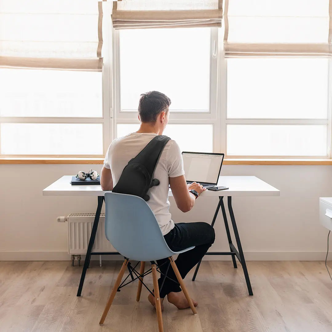 Person using Green Lion Neck & Shoulder Massagers, sitting at a desk with a laptop in a bright room with large windows.