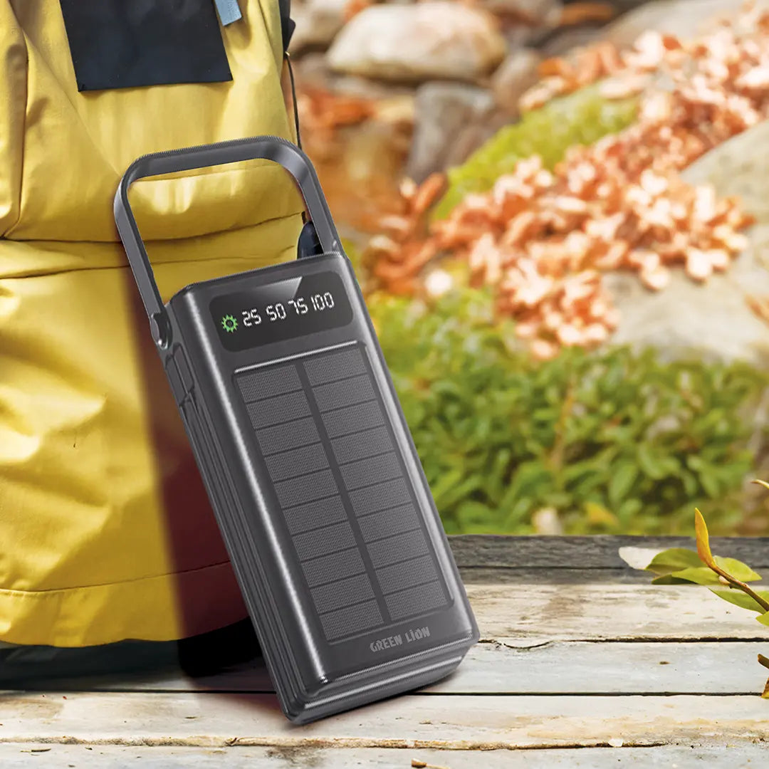 A Green Lion solar power bank with a built-in handle and digital display, placed outdoors near a backpack, highlighting its portability for travel and outdoor use.