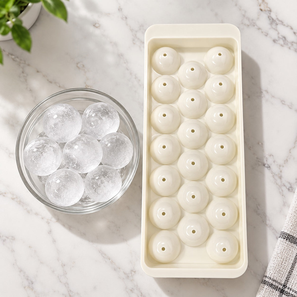 A rectangular ice mold tray filled with round ice balls, placed on a white marble surface next to a glass bowl of clear spherical ice. A small plant leaf and folded cloth are visible at the edges.