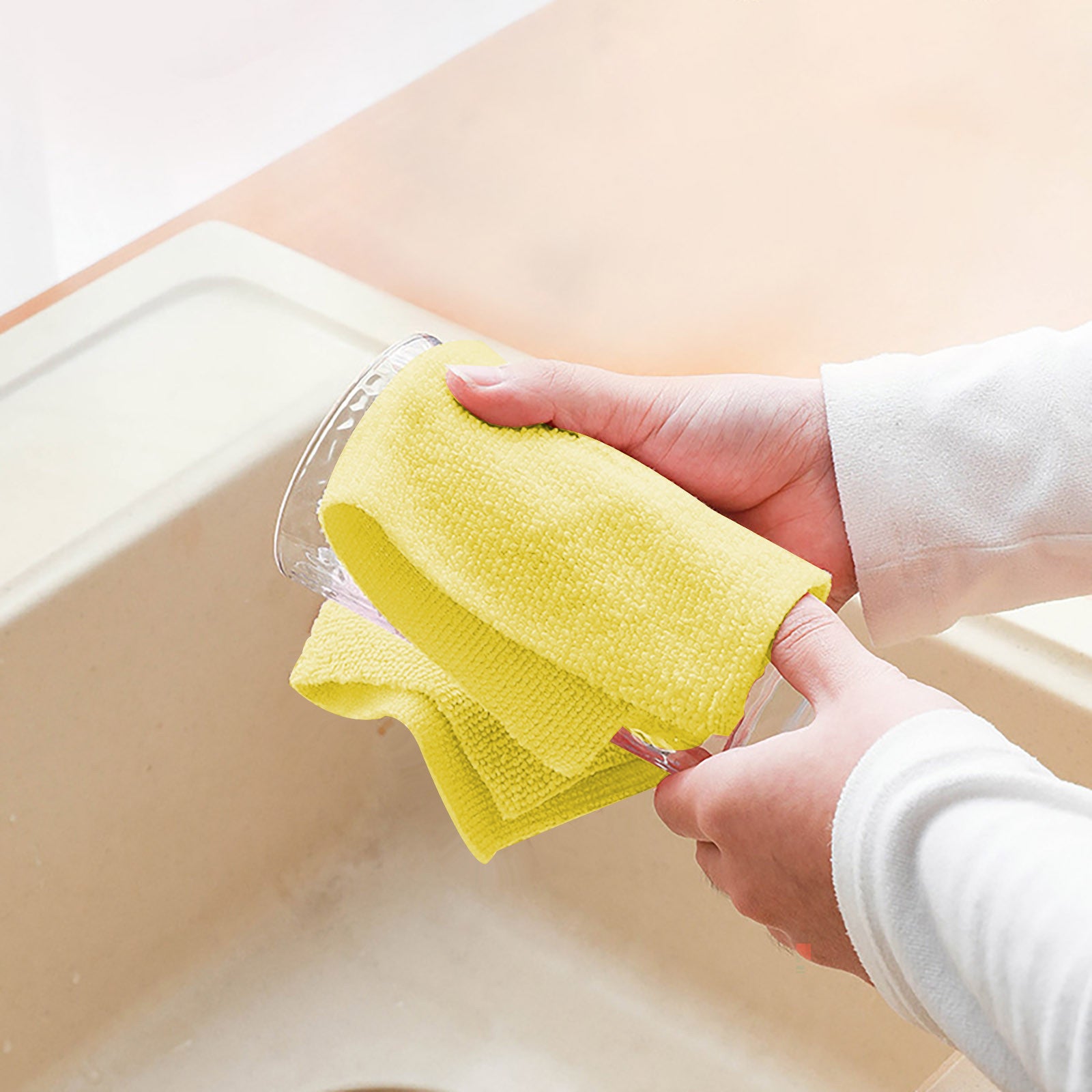 A person's hands holding a clear glass cup while wiping it with a bright yellow Reusable Multipurpose Cleaning Cloth Roll in a beige kitchen sink.