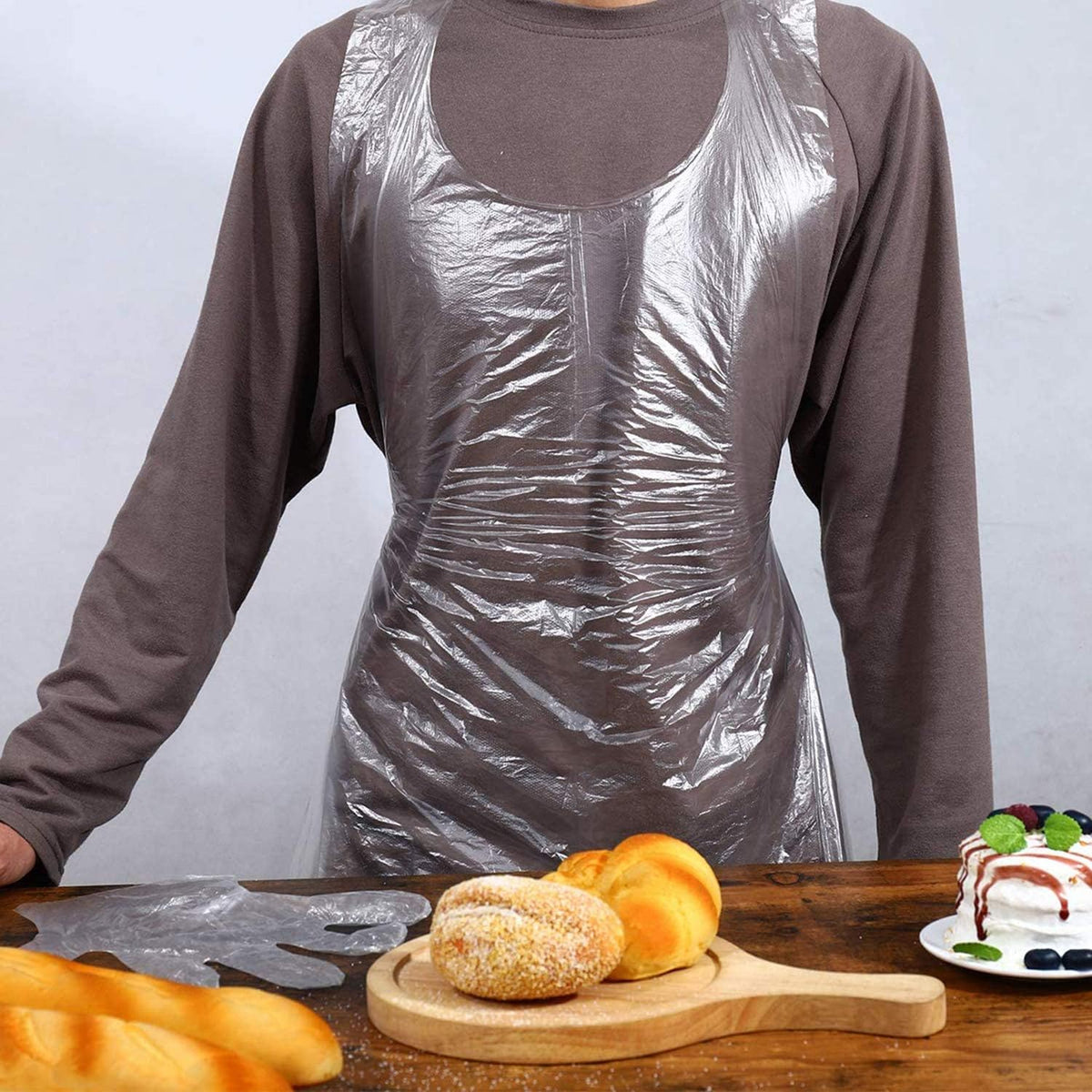 Person wearing a plastic apron standing behind a table with bread and a cake.