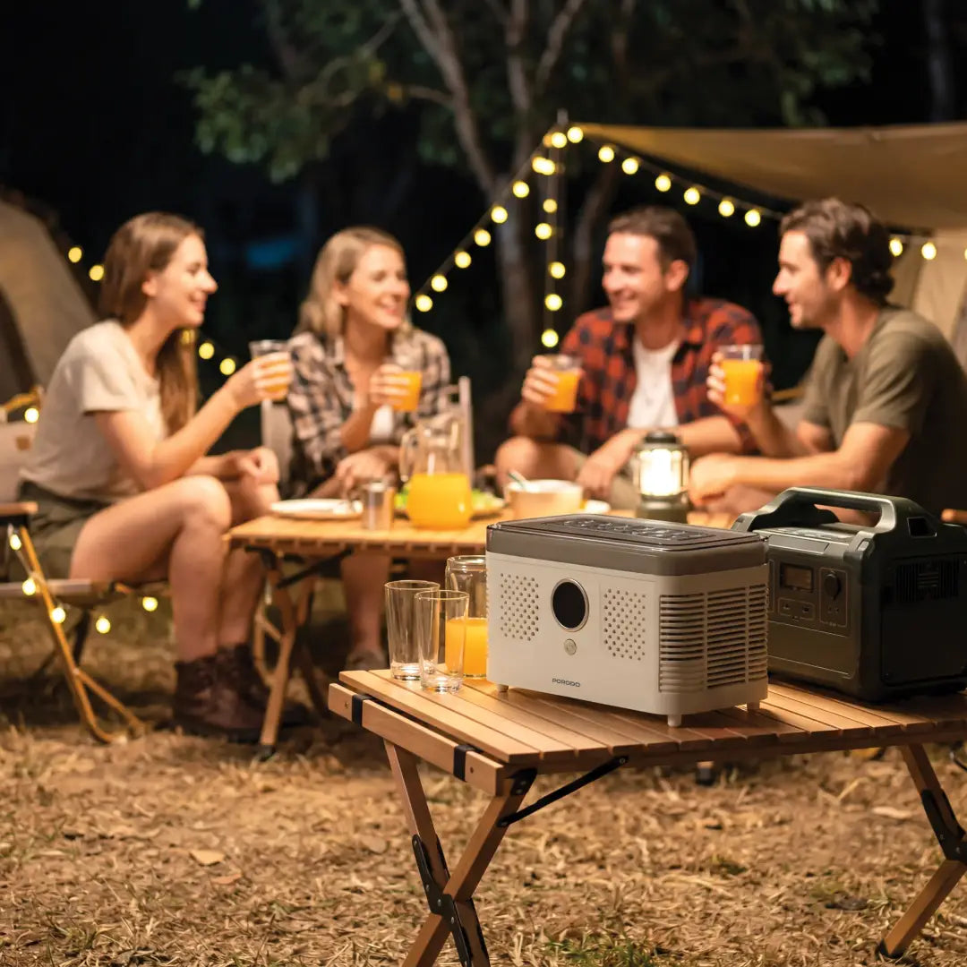 A PORODO FrostIQ Portable Ice Cube Maker and power station set on a folding camp table at an outdoor night campsite, with four friends laughing and drinking orange juice in the background under string lights.