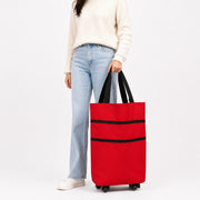 A person wearing a cream sweater, light blue jeans, and white sneakers holds a bright red rolling Shopping Trolley Bag gwith black handles and black horizontal trim against a clean white studio background.