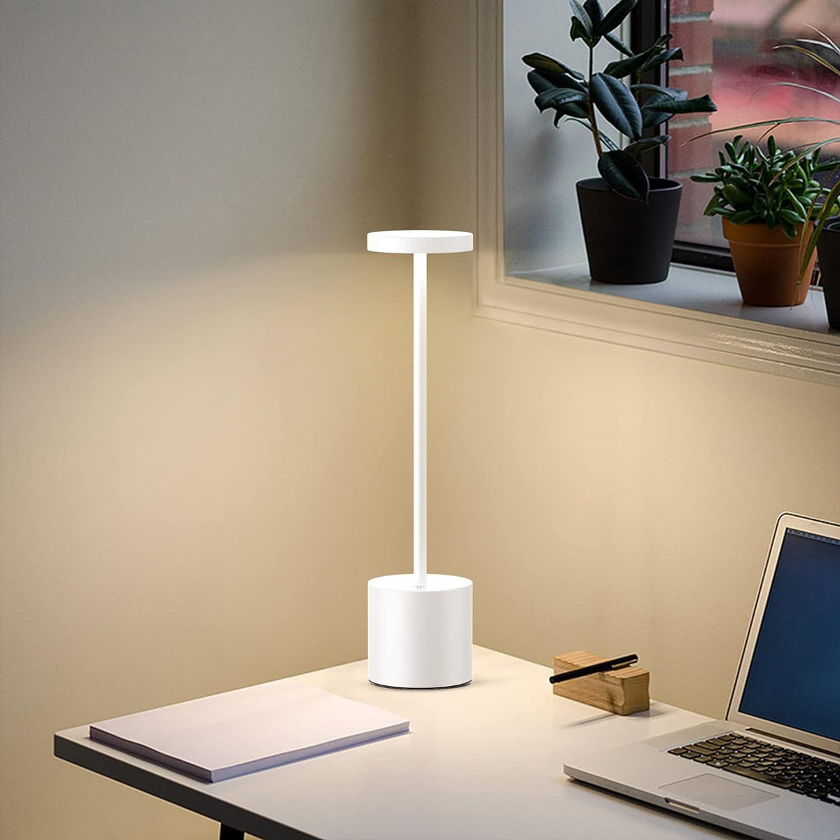 White desk lamp on a desk with a laptop and notebook, near a window with plants.