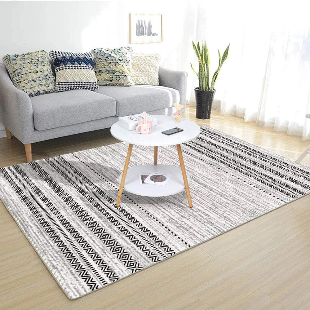 A round white side table is placed on a patterned rug in a bright, cozy living room featuring a grey sofa with textured cushions and an indoor plant by the window