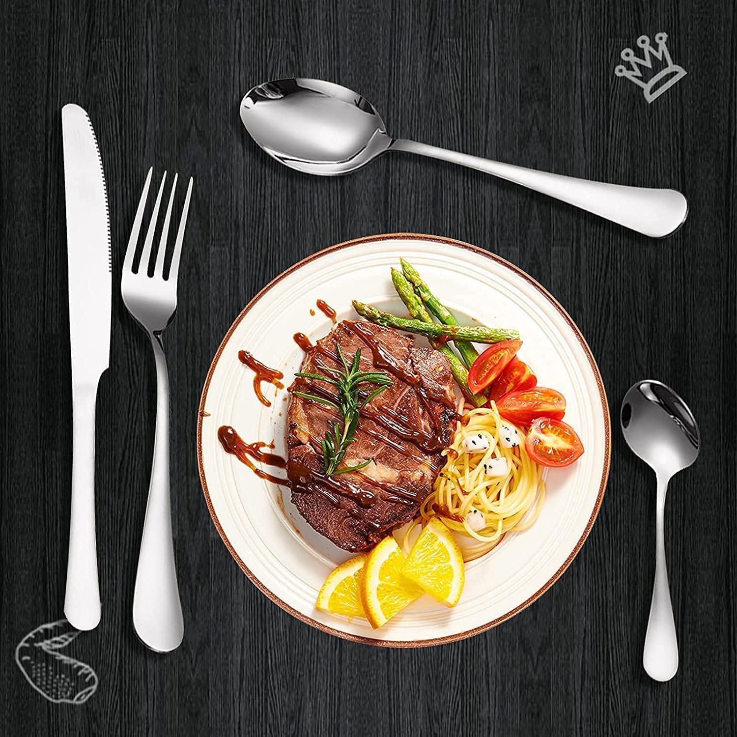 Elegant stainless steel 24-piece cutlery set arranged with a knife, fork, and two spoons beside a plated steak meal with pasta, vegetables, and lemon slices on a dark tabletop.