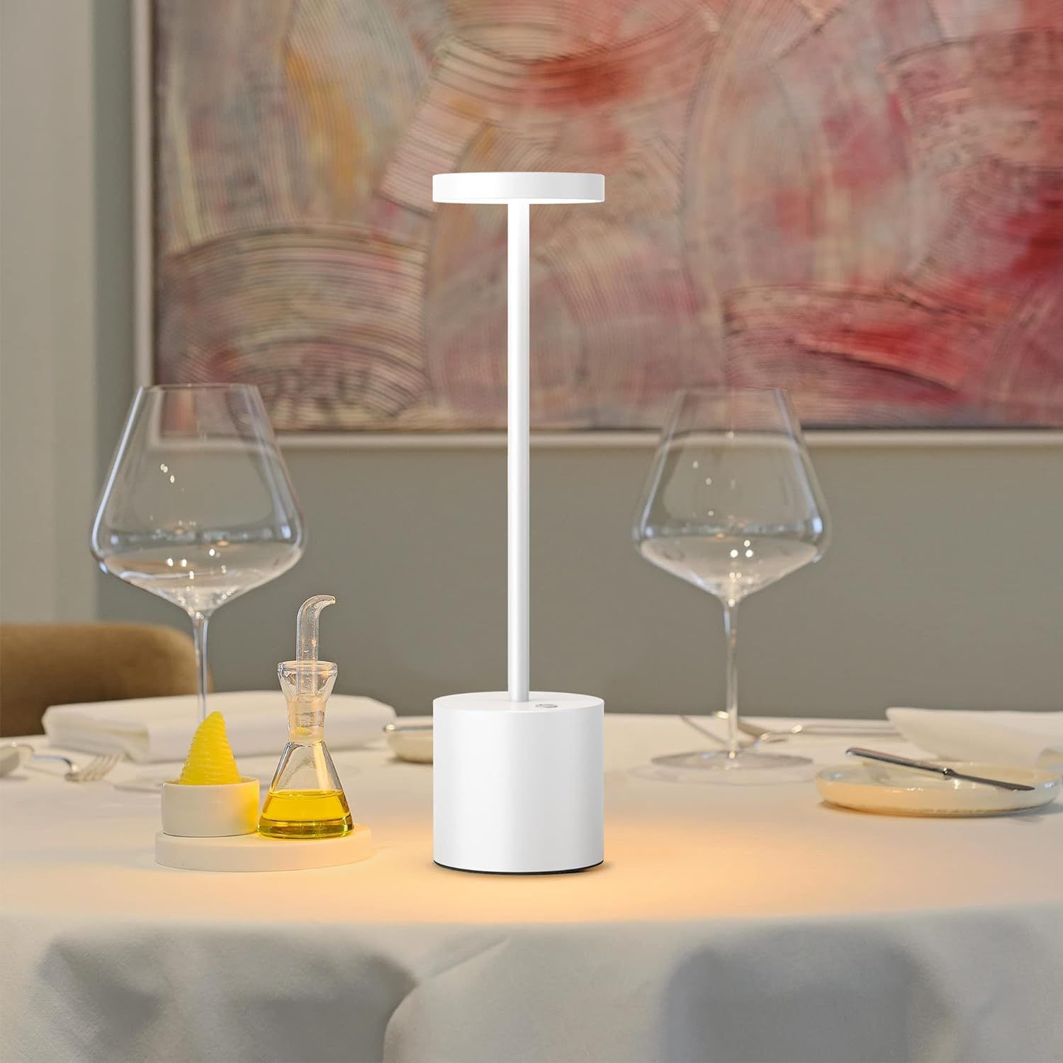 White lamp on a table with wine glasses and a painting in the background