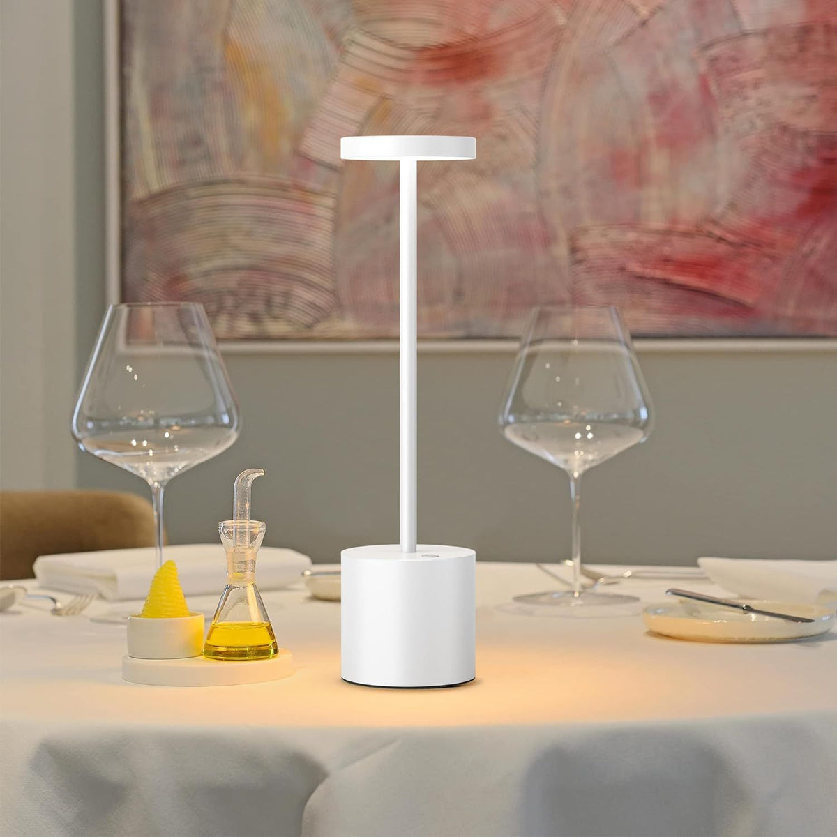 White lamp on a table with wine glasses and a painting in the background