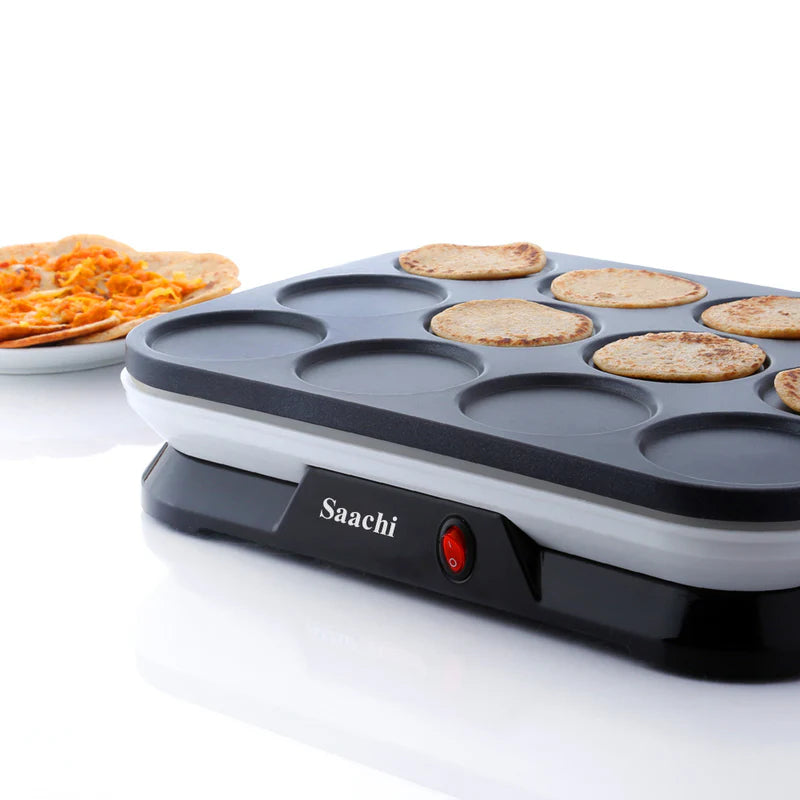 An electric mini pancake maker with a non-stick surface from Saachi, cooking several pancakes at once. A plate of cooked pancakes is placed nearby on a white surface