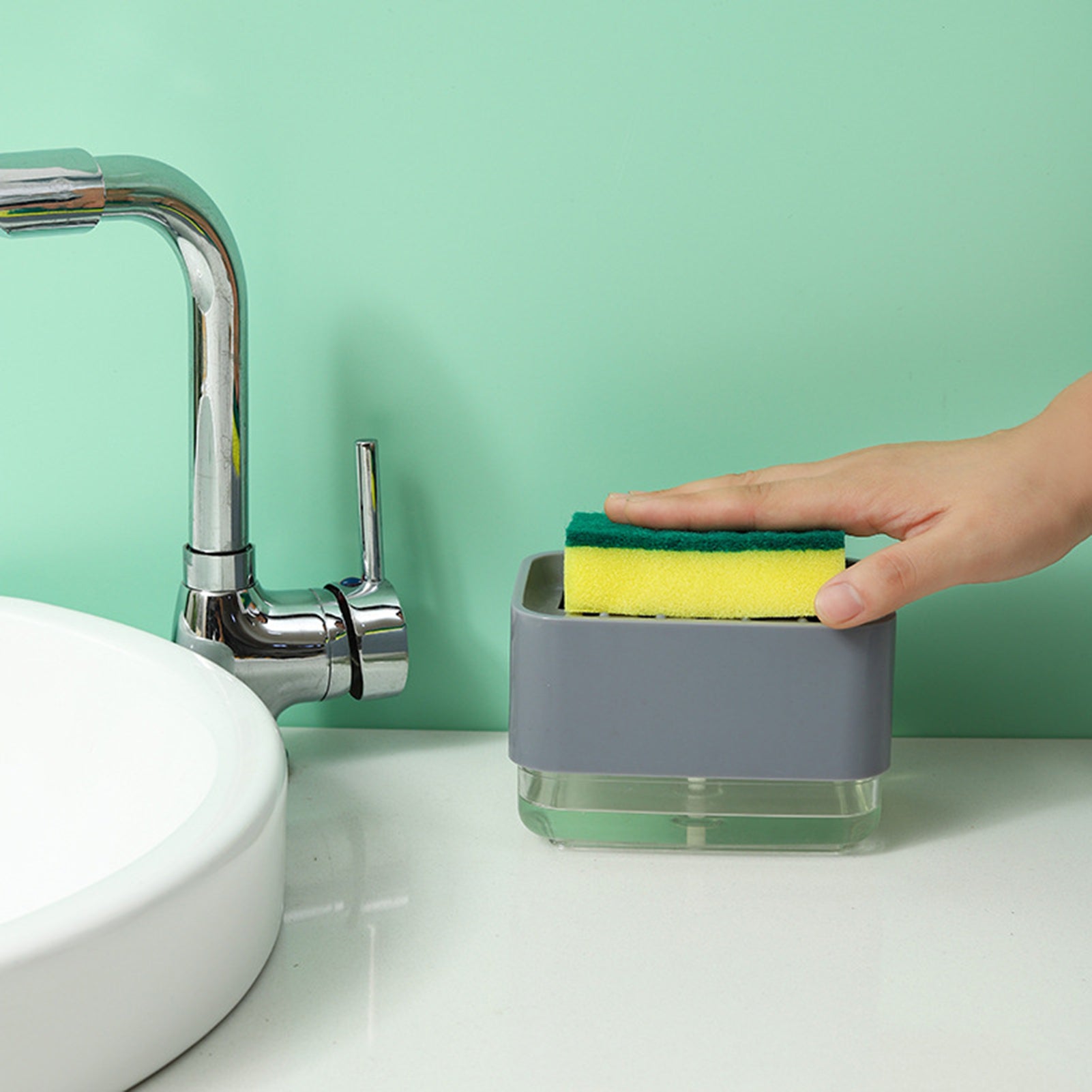 A compact sponge holder with a built-in soap dispenser, shown being pressed with a sponge to release liquid soap, placed beside a kitchen sink for easy dishwashing.