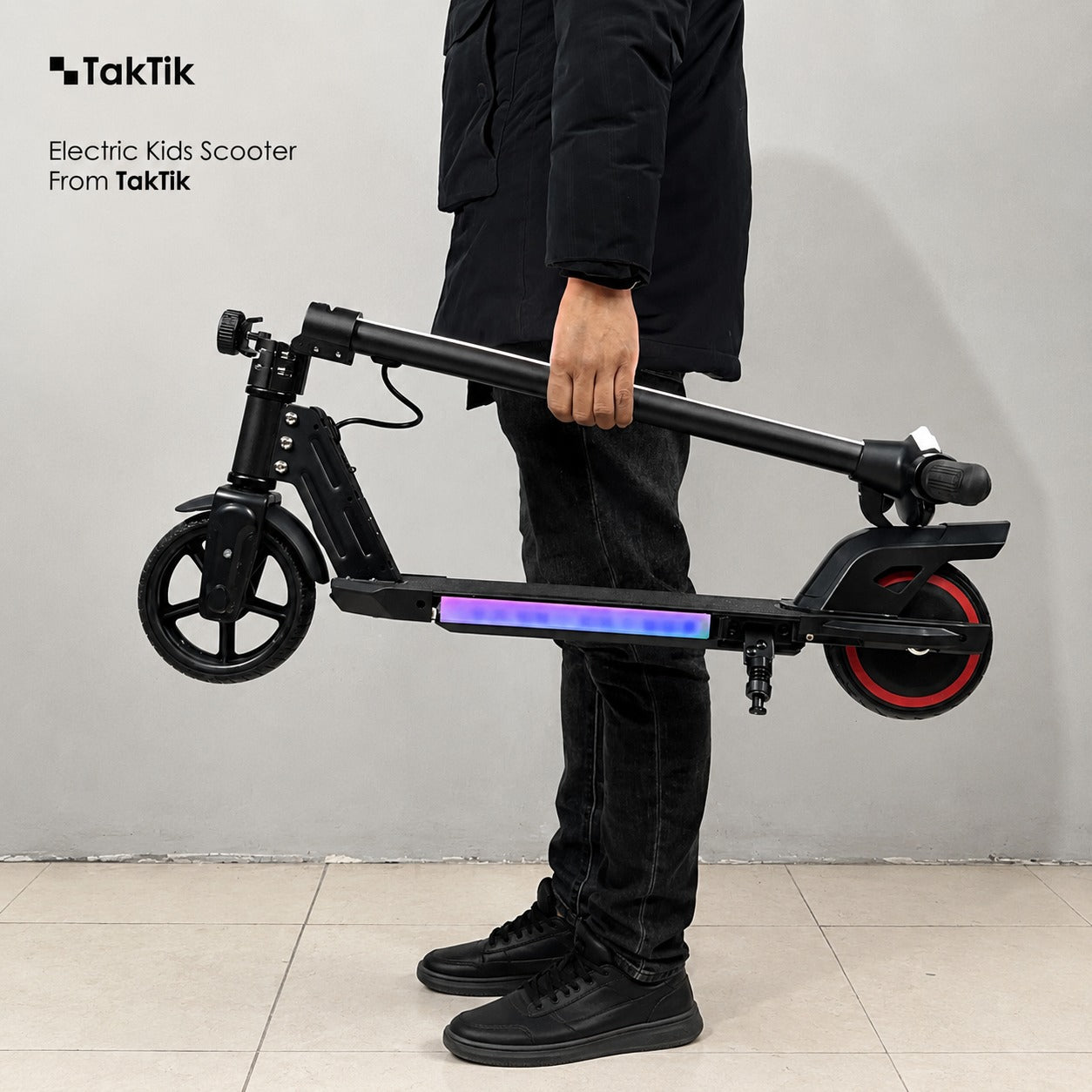 A person in a black puffer jacket, jeans, and black sneakers holds a folded black TakTik Kids Electric Scooter with illuminated purple and blue LED striping on the deck. The scooter has a black frame, black wheels, and a red accent ring around the rear wheel.