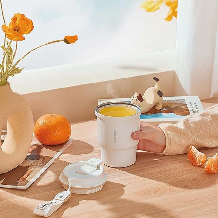 A hand holds a white Lepresso Thermo Brew Insulated Tumbler filled with a yellow liquid on the left, a light-colored, curved ceramic vase holds bright orange poppy flowers.