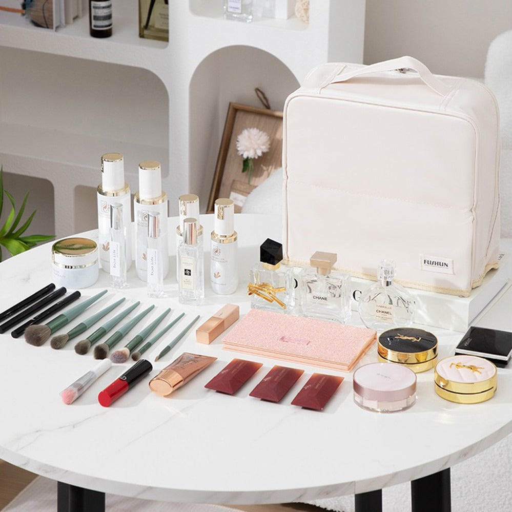 A Travel Makeup Bag Cosmetic Organizer Case with a top handle sits on a white marble table, surrounded by an assortment of cosmetics including brushes, lipsticks, palettes, and perfume bottles.