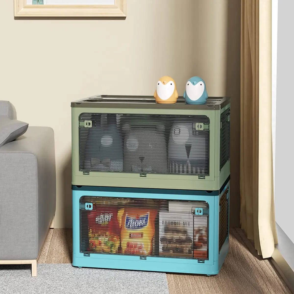 2 Foldable Transparent Storage Box with Wheels 1 in green and 1 in blue kept in a room next to a sofa