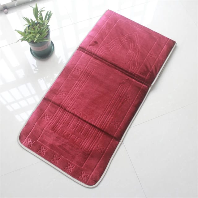 Red color Islamic Foldable Prayer Mat kept flat on a white surface next to a plant 